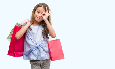 Brunette hispanic girl holding shopping bags with happy face smiling doing ok sign with hand on eye looking through fingers