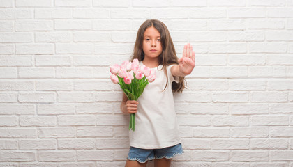 Young hispanic kid over white brick wall holding flowers on mother day with open hand doing stop sign with serious and confident expression, defense gesture