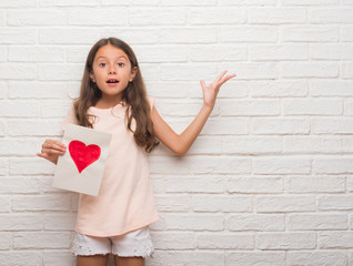 Young hispanic kid over white brick wall giving mother day card very happy and excited, winner expression celebrating victory screaming with big smile and raised hands