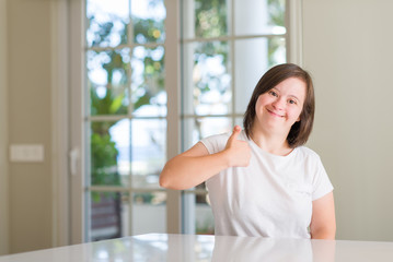 Down syndrome woman at home doing happy thumbs up gesture with hand. Approving expression looking at the camera with showing success.