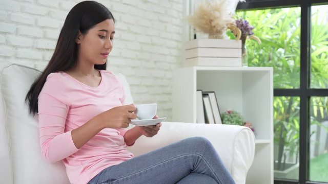 Cheerful asian young woman drinking warm coffee or tea enjoying it while sitting in her living room at home. Attractive happy asian woman holding a cup of coffee.