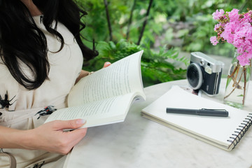 Woman reading a book in a cafe with white notepad black pen camera glass vase pink flower and green background