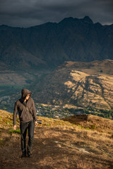 Young male photographer walking on Queenstown hill with mountain scenery in the background during golden hour sunset in Queenstown, South Island, New Zealand. Travel and photography concepts