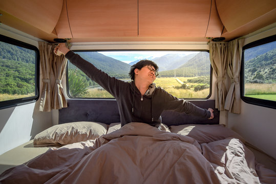 Young Asian man stretching arms while staying in the blanket in camper van in the morning with mountain scenery in the background. Road trip in summer of South Island, New Zealand.