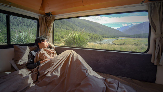 Young Asian man staying in the blanket looking at mountain scenery through the window in camper van in the morning. Road trip in summer of South Island, New Zealand.