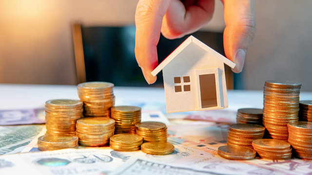 Real estate or property investment growing business. Home mortgage loan rate. Saving money for retirement concept. male hand holding house model over coin stack on international banknotes on table
