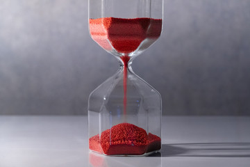Red sand running through the shape of modern hourglass on white table.Time passing and running out...