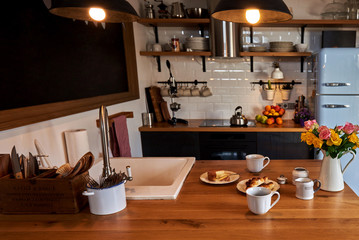 Rural breakfast homemade cheese and croissants with roses bouquet in jug. Kitchen in the attic and rustic style.  Wall with shelves, trays, jars, mugs, sink. In the background a wall of white brick.