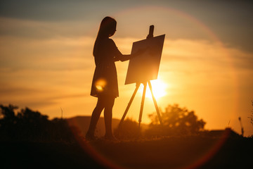 Obraz premium Silhouette of a blonde girl artist. Lady paints a painting on the canvas with the help of paints. A wooden easel keeps the picture. Summer is a sunny day, sunset