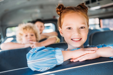 close-up portrait of smiling little schoolgirl riding on school bus with classmates behind
