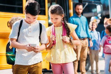 adorable schoolgirl and schoolboy using smartphone together in front of school bus with classmates