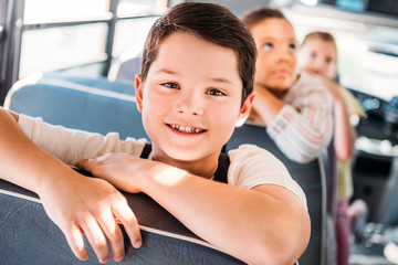 close-up portrait of smiling little schoolboy riding on school bus with blurred classmates on...