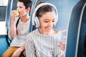 happy little schoolgirl using smartphone and listening music with headphones while riding on school bus