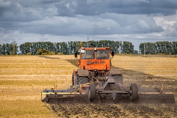 An orange modern tractor plows the earth in.  a golden field of wheat on a summer day, in the sky a cumulus cloud, in the background a forest.
