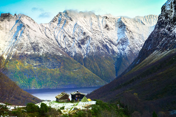 Group of hikers walking in the mountains. A small wooden cabin, tents and holiday makers in the alley in the middle of mountains, next to Fjords. End of autumn, cold weather, bonfire and snowy peaks
