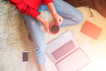 Top view of young woman sitting on floor with laptop. Young woman dressed in red pullover sitting on floor and holding laptop while using smartphone. Online shopping. Girl chooses gifts online.