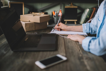 Close up of a female hand writing on an blank notebook with a pen. Side view of woman's hands. She is using his latpot and writing numbers down. Concept of accounting work. Selective focus.