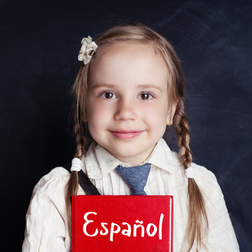 Kid learning spanish in language school. Happy little girl holding book in her hands on blackboard background