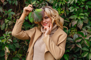 Young beautiful woman in sweater and coat stands next to the background of wild grapes leaves