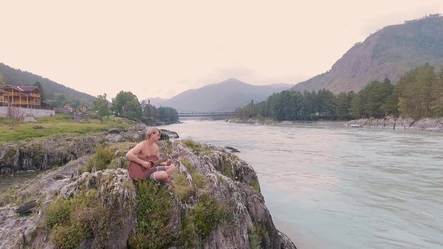 A man sitting on the rocks by a mountain river and playing acoustic guitar. aerial survey. 4k