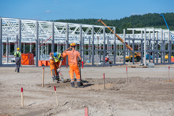 Industrial construction site of new commercial shopping mall. Workers in front, concrete piles foundation for the building, cranes and trucks in background. Ongoing framing, construction of factory