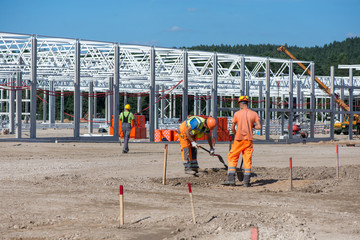 Industrial construction site of new commercial shopping mall. Workers in front, concrete piles foundation for the building, cranes and trucks in background. Ongoing framing, construction of factory