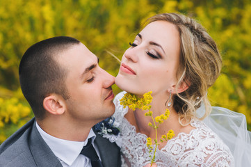 A stylish bridegger strokes a beautiful bride with curly hair in a lace dress on a plaid. Wedding portrait close-up of a happy newlyweds in nature with yellow flowers.