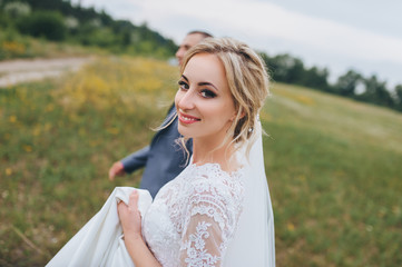 Beautiful newlyweds go to the yellow field in a coniferous forest. The bride in a lace dress smiles. Wedding portrait of happy newlyweds.