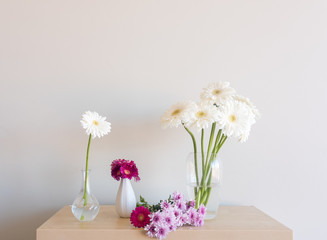White and pink gerberas and purple chrysanthemums in vases and on wooden shelf against neutral wall background (selective focus