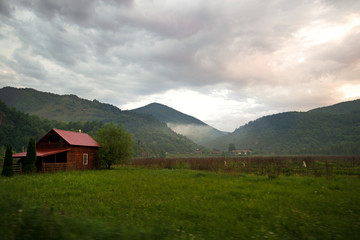 A village house built in meadow in front of mountains. Small cloud formed from the early morning mountain dew evaporating on a hot day. Sun is rising behind mountains