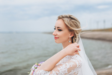 Fototapeta na wymiar A beautiful bride in a lace dress dreams at the pier, against the background of the sea and sky. Wedding portrait close-up of a curly bride on the waterfront.