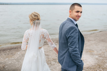 A beautiful bride in a lace dress and a stylish groom stand on the pier, against the background of the sea and sky. Wedding portrait of lovely newlyweds. Wedding day.