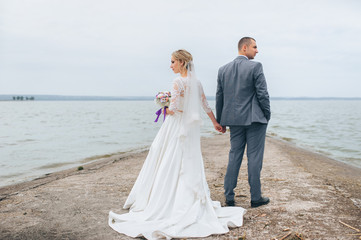 Fototapeta na wymiar A beautiful bride in a lace dress and a stylish groom stand on the pier, against the background of the sea and sky. Wedding portrait of lovely newlyweds. Wedding day.