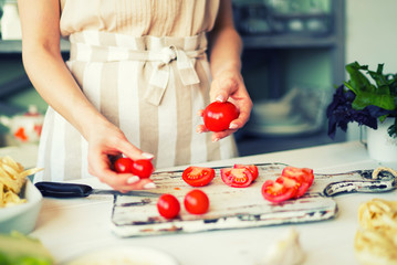 Young woman in apron at table with vegetables. Сooking meal at home, preparing lunch from fresh organic vegetables in light kitchen with wooden surface, full of rural kitchenware. Selective focus.