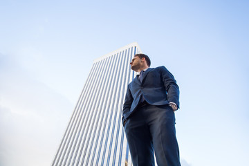 From below view of stylish man in elegant suit posing confidently on background of modern office skyscraper
