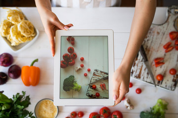 Hands with the tablet computer  pictures of food. Various fresh Ingredients for cooking tasty vegetarian lunch from fresh vegetables. Sliced vegetables prepared for making meal. Selective focus.