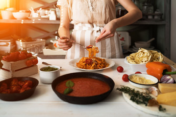 Woman in kitchen making delicious pasta with sauce using a pan at home. Italian rural cooking still life. Wooden board, fresh vegetables, cooked pasta and a pan of sauce on a wooden kitchen table.
