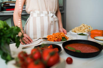  Woman in kitchen making delicious pasta with sauce using a pan at home. Italian rural cooking still life. Wooden board, fresh vegetables, cooked pasta and a pan of sauce on a wooden kitchen table.