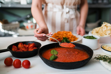 Woman in kitchen making delicious pasta with sauce using a pan at home. Italian rural cooking still life. Wooden board, fresh vegetables, cooked pasta and a pan of sauce on a wooden kitchen table.