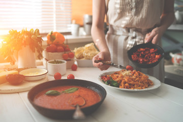 Young woman in kitchen making delicious pasta with sauce using a pan at home. Italian rural cooking still life. Wooden board, fresh vegetables, cooked pasta and a pan of sauce on wooden kitchen table.