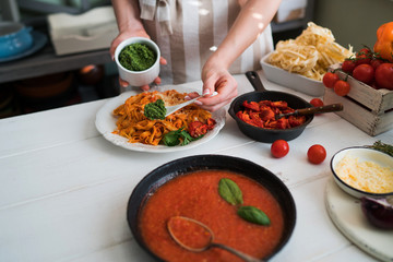 Young woman in kitchen making delicious pasta with sauce using a pan at home. Italian rural cooking still life. Wooden board, fresh vegetables, cooked pasta and a pan of sauce on wooden kitchen table.