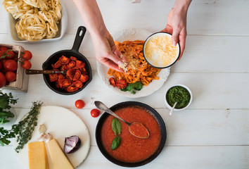 Cook's hand sprinkling chopped cheese in the tomato sauce pasta on the table. Tomatoes, garlic, parsley, pepper, and parmesan cheese on white wooden background. Selective focus. Top view