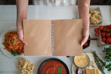 Young woman is holding cookbook and writing a new recipe. Female chef with cookbook. Cooking, culinary, food and people concept. Fresh organic vegetables and spices on a wooden background. Heatly food