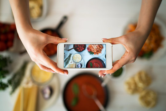 Hands with the smart phone pictures of meal. Young woman, cooking blogger is cooking at the home kitchen in sunny day and is making photo at smartphone. Instagram food blogger workshop concept.