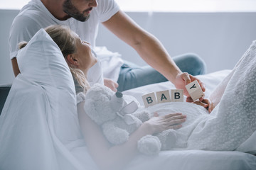 Obraz premium partial view of man putting wooden blocks with baby lettering on belly of pregnant wife with teddy bear at home