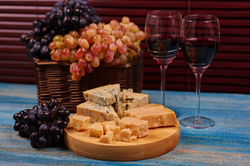 Light and dark blue organic tasty fruit arranged in wicker basket standing on vintage table near to pieces of moldy cheese and couple wine glasses.