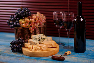 Colorful bunches of grape arranged in wicker basket standing on blue retro table near to bottle of tasteful red wine and cheese for degustation.