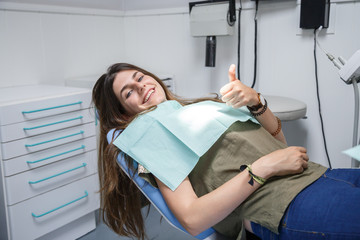 Portrait of a blue-eyed young woman sitting in the chair of a dental clinic gesturing thumb up 