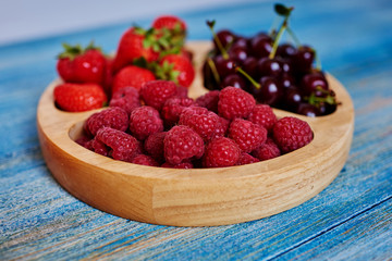 Fresh strawberries, raspberries and cherries are in a beautiful wooden cookware on the table, vintage blue