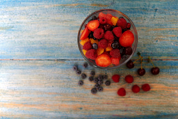 Top view on a vintage wooden desk with peeling paint, there is a deep glass bowl with fresh chopped salad of ripe berries